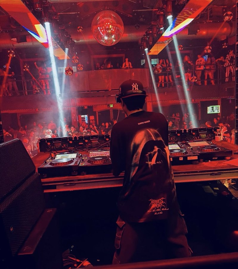 The floor seen from behind the decks: a packed two-level room under red light and beams.